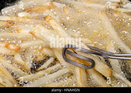 Medium sized Russet potatoes peeled, sliced and frying in hot oil creating home made french fries, stirring while cooking with tongs. An inexpensive a Stock Photo