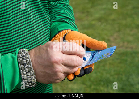 Male hand with golf score card keeping score writing down score with green grass of golf course in the background. Wearing a survival bracelet. Stock Photo