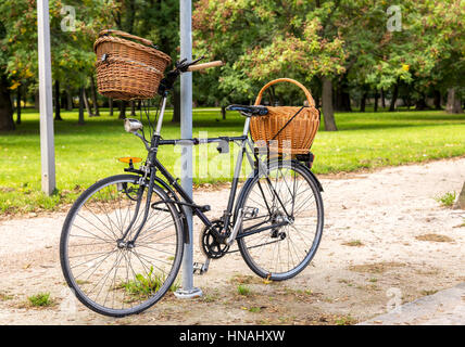 A black bicycle with two wickers baskets leans against a post on a gravel path. Stock Photo