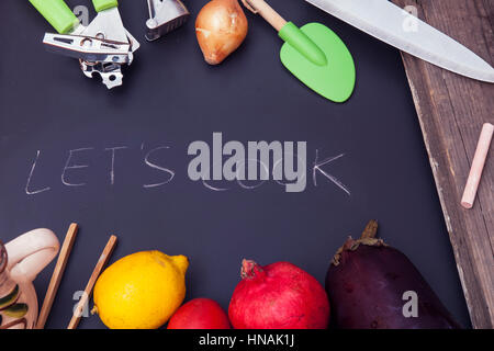 Let’s cook text written with white chalk on black chalkboard with kitchen tools and vegetables around Stock Photo