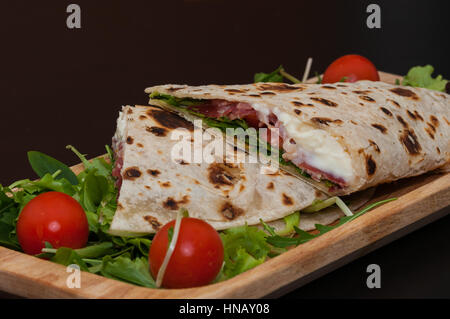 italian piadina, with salad, ham, cheese, mozzarella and tomatoes, wood dish and black background Stock Photo