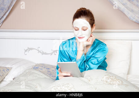 Beautiful woman with green nightie and white facial mask resting on bed and holding tablet before sleeping. healthcare and technology. Stock Photo
