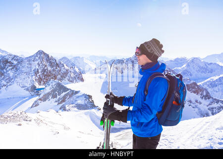 Skier standing with ski in hand enjoying the panoramic landscape view of the snow covered Alps mountains resort during a sunny day Stock Photo