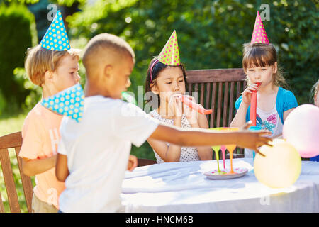 Kids celebrate friend's birthday in garden in summer with noise makers and hats Stock Photo