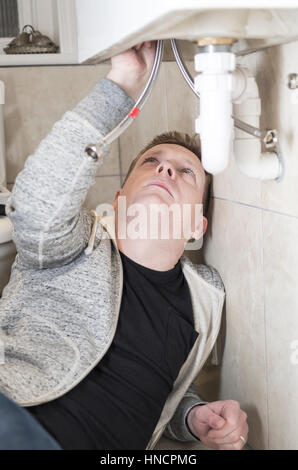 Young man attempting Do It Yourself (DIY) plumbing at home under a sink fitting a new tap looking upwards. Stock Photo