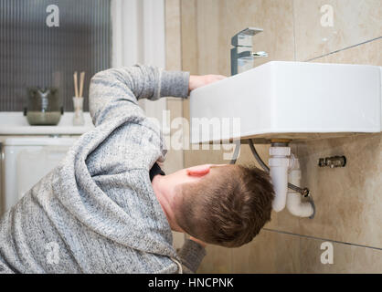 Rear view of a young man attempting Do It Yourself (DIY) plumbing at home under a sink fitting a new tap. Stock Photo
