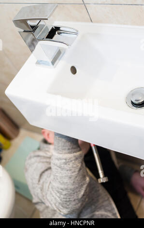 Young man attempting Do It Yourself (DIY) plumbing at home under a sink fitting a new tap with head obscured Stock Photo