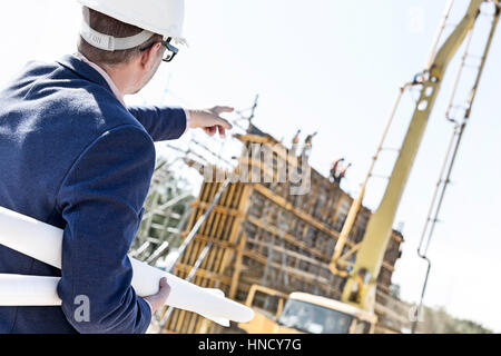 Rear view of architect holding blueprints while pointing at construction site Stock Photo