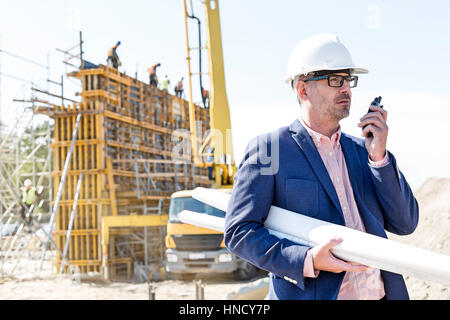 Architect using walkie-talkie while holding blueprints at construction site Stock Photo
