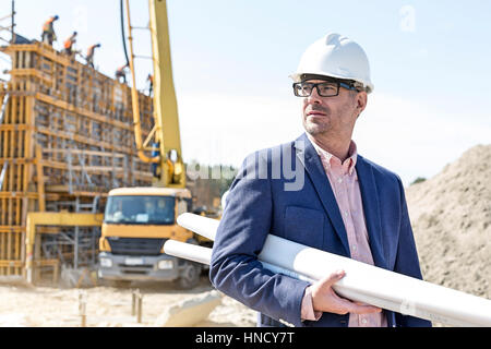 Confident architect holding rolled up blueprints at construction site Stock Photo