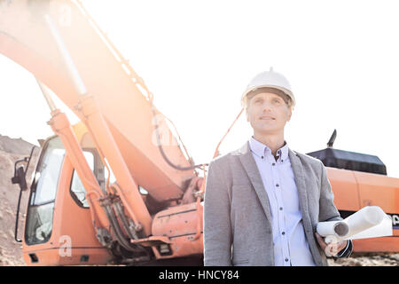 Thoughtful engineer looking away while holding blueprints by bulldozer at construction site Stock Photo