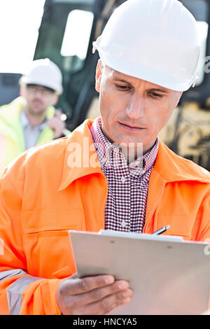 Supervisor writing on clipboard at construction site with colleague in background Stock Photo