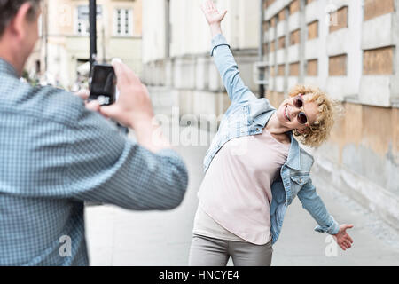 Man photographing playful woman standing with arms outstretched on city street Stock Photo