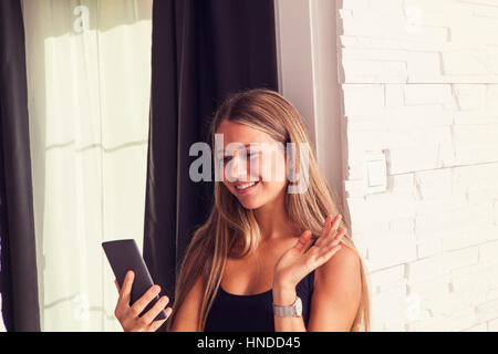 Pretty blonde girl in black shirt near the window making a video call with smart phone, saying hello with the hand smiling at the screen Stock Photo
