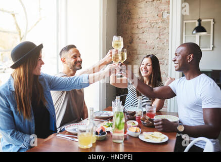 Four young adult sitting at table and clinking glasses of wine while having dinner together in restaurant. Stock Photo