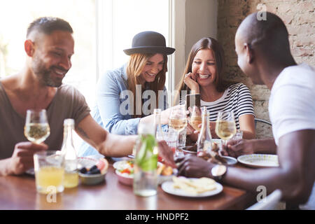 Group of four young friends having dinner in cozy cafe while women sharing new on phone. Stock Photo