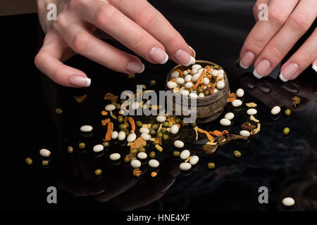 Female hands with beautiful French manicure gather the haricot scattered on a black background, peas, dried vegetables and spices in a vintage melkhiorovy saltcellar Stock Photo