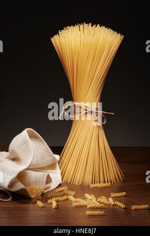 Dry spaghetti pasta tied up with rope and fusilli pasta falling out of burlap bag on dark wooden table on grey background Stock Photo
