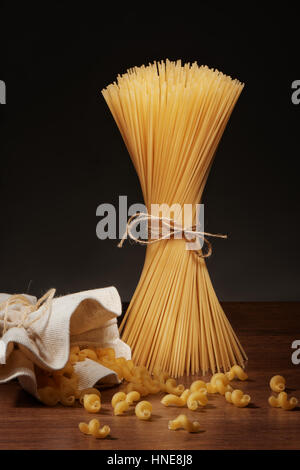 Dry spaghetti pasta tied up with rope and cavatappi pasta falling out of burlap bag on dark wooden table on grey background Stock Photo