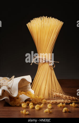 Dry spaghetti pasta tied up with rope, cavatappi pasta falling out of burlap bag and wheat ears on dark wooden table on grey background Stock Photo