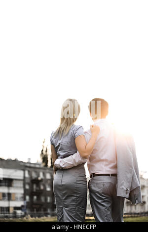 Rear view of business couple standing with arms around against clear sky on sunny day Stock Photo