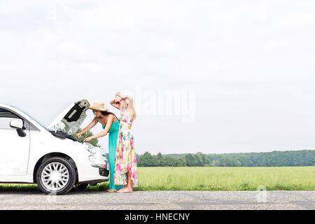 Female friends examining broken down car on country road against clear sky Stock Photo