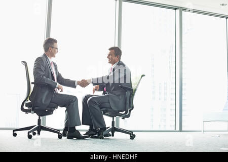 Full-length side view of businessmen shaking hands while sitting on office chairs by window Stock Photo