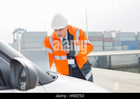 Middle-aged man talking on walkie-talkie while using laptop in shipping yard Stock Photo