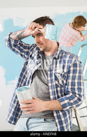 Tired man holding paint can and brush with woman painting in background Stock Photo