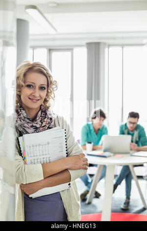 Portrait of beautiful businesswoman holding files with colleagues working in background at creative office Stock Photo