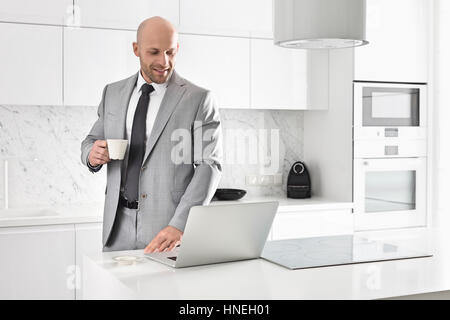 Mid adult businessman having coffee while using laptop in kitchen Stock Photo