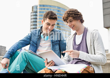 Happy young male college students using digital tablet against building Stock Photo