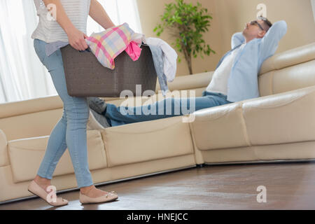 Low section of woman walking with laundry basket while man relaxing on sofa in background Stock Photo