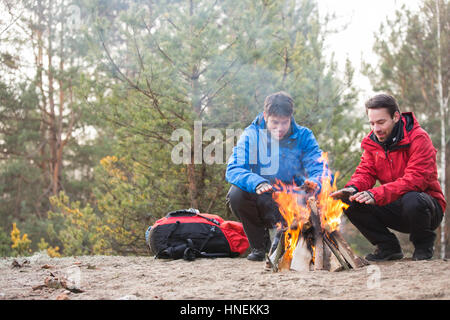 Male backpackers warming hands at campfire in forest Stock Photo