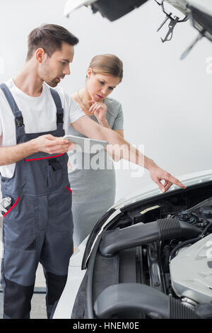 Young repair worker explaining car engine to worried customer in workshop Stock Photo