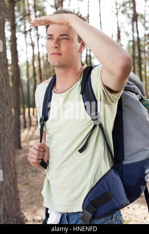 Young man with backpack shielding eyes in forest
