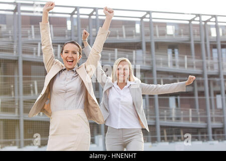 Excited young businesswomen with arms raised against office building Stock Photo