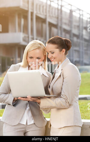 Happy young businesswomen using laptop together against building Stock Photo