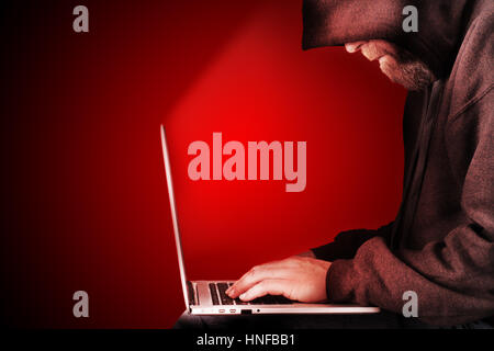 Male computer hacker wearing a hooded top leaning over a laptop with red warning background. The screen light illuminates the man with a beard perform Stock Photo