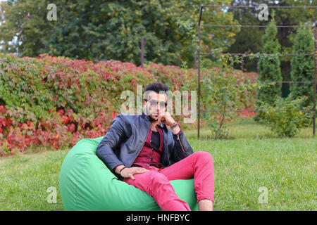Arabian Young Man Sitting and Resting From City Bustle in Soft Green Chair, smiles and Looks Around. Handsome Guy Has Dark Hair and Black Beard and dr Stock Photo