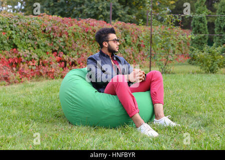 Arabian Young Man Sitting and Resting From City Bustle in Soft Green Chair, smiles and Looks Around. Handsome Guy Has Dark Hair and Black Beard and dr Stock Photo