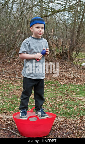 This Caucasian 5 year old boy is playing outside and standing on a red bucket that is frozen solid ice.  He's wearing a blue stocking cap, black sweat Stock Photo