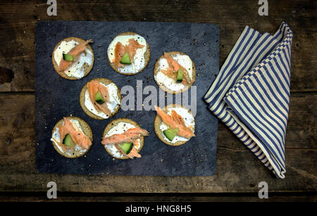 Oat Cake Cucumber and Salmon canapés on a black slate and rustic dark wood background with cracked black pepper and a blue striped napkin Stock Photo