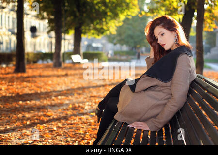 Beautiful red haired woman on a bench at the city park. Seasonal shot Stock Photo