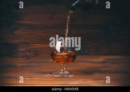 Pouring brandy or cognac from the bottle into the glass against wooden background Stock Photo