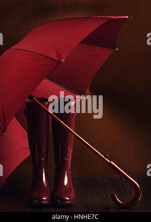 Photo in low key. Rubber boots and umbrella burgundy color on a brown background. Selective focus. Stock Photo