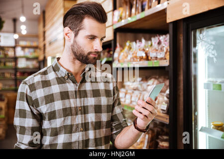 Handsome bearded young man in plaid shirt using cell phone in grocery shop Stock Photo