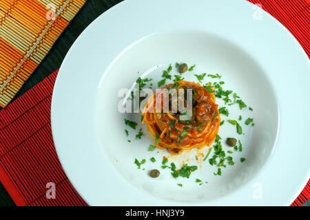 Spaghetti puttanesca on colorful placemats lying on wooden table - Italian recipe with garlic, olive oil, anchovies, olives, tomato, parsley, capers Stock Photo