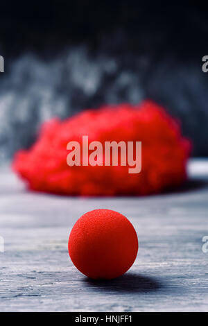 a curly red hair wig and a red clown nose forming the face of a clown on a rustic wooden surface Stock Photo