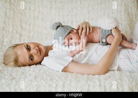 Baby boy wearing a rabbit costume, hugged by his mother that is sitting on her back on a bed, contemplating Stock Photo